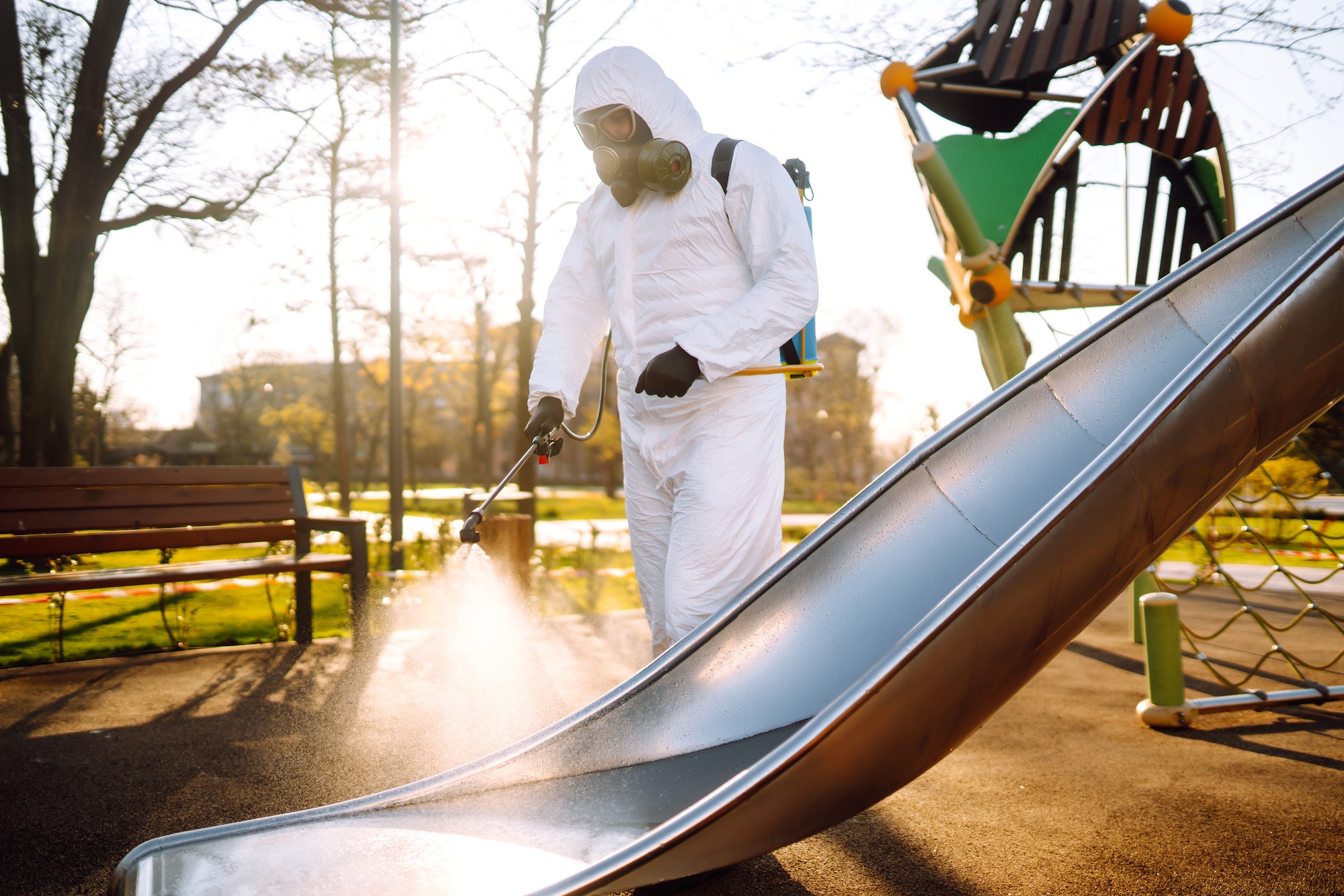 Man wearing protective suit disinfecting the playground in the sun with spray. Covid -19. Cleaning.